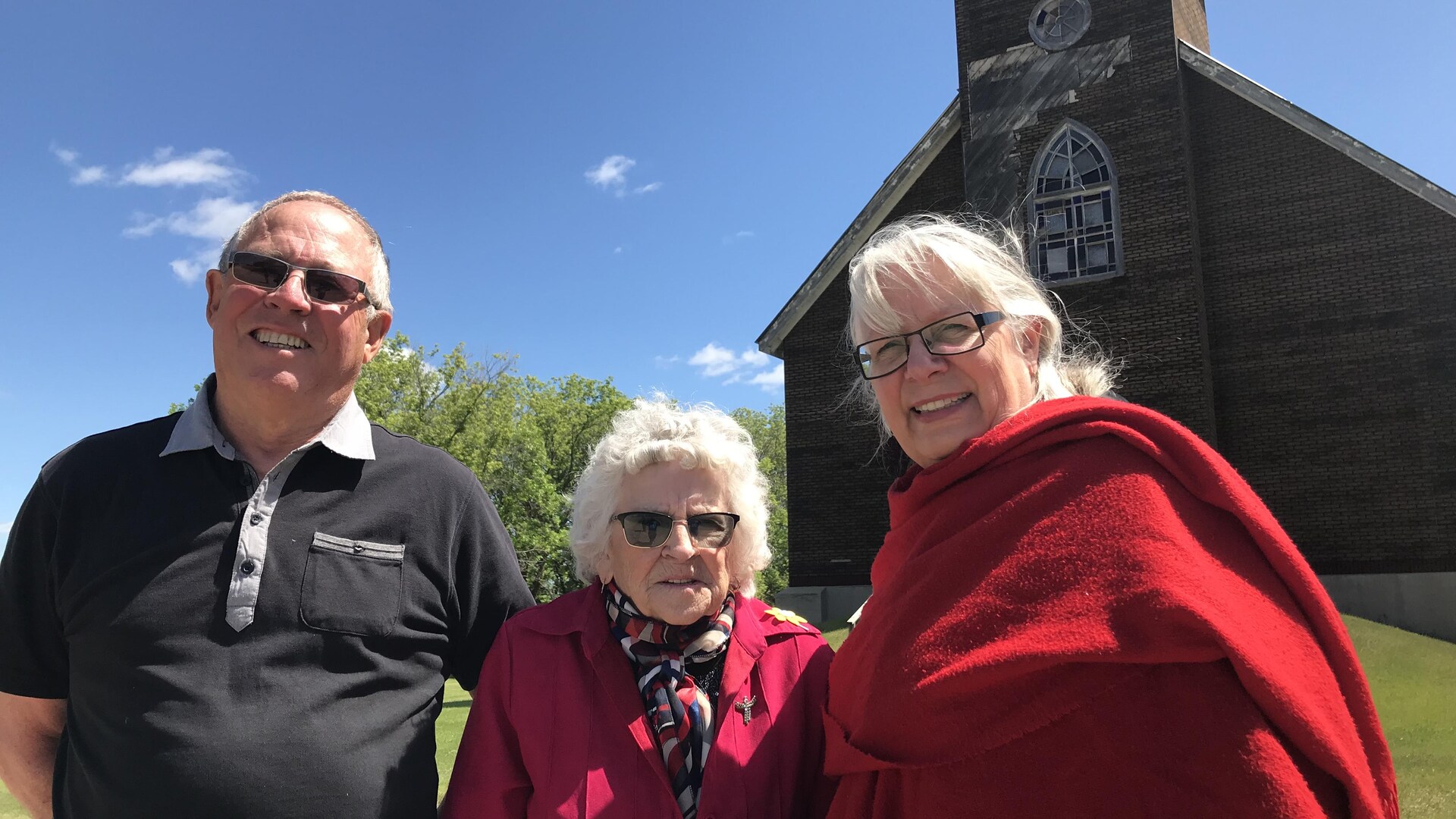 Trois personnes posent en souriant devant l'église Sainte-Amélie au Manitoba. 