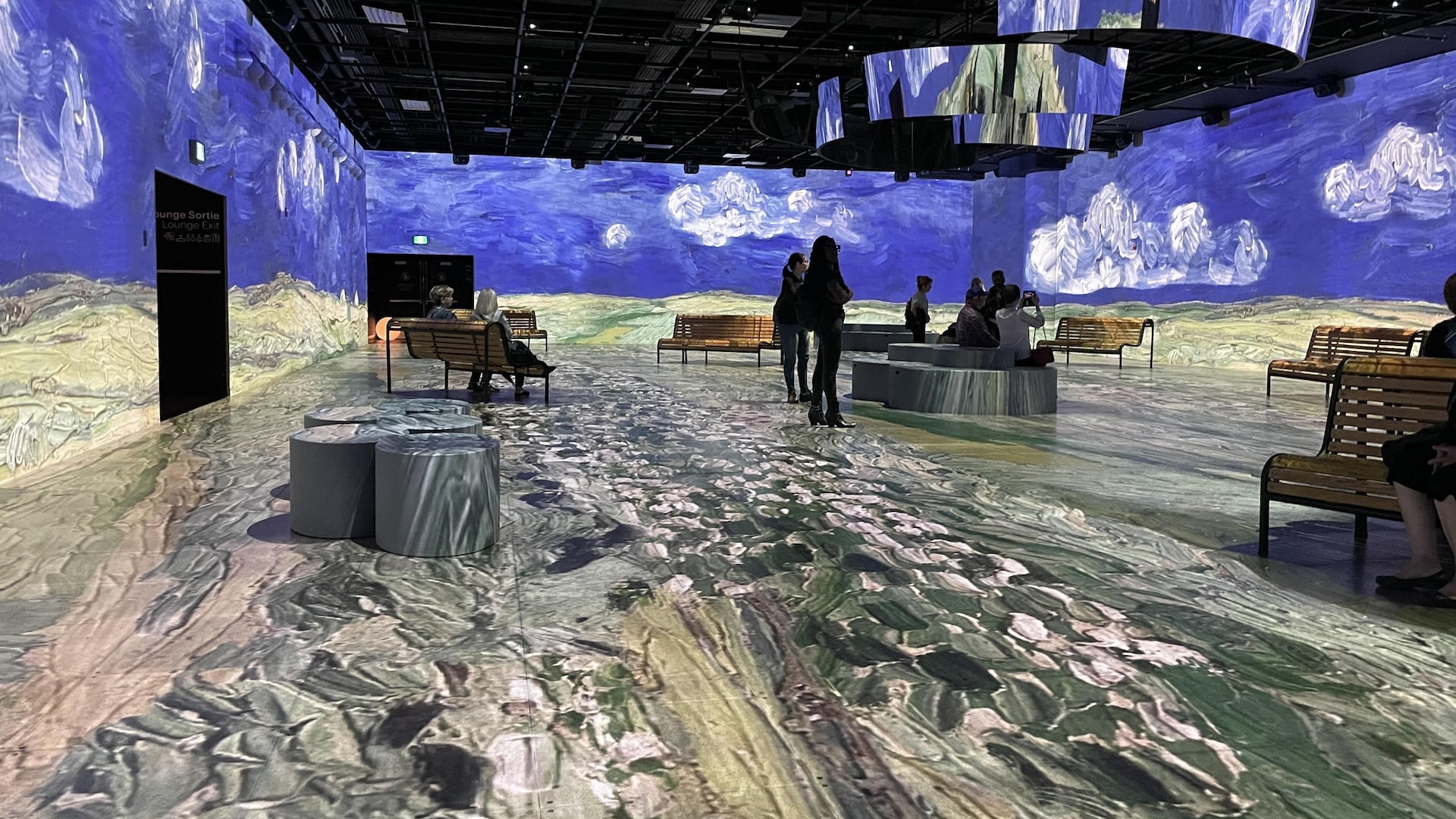 Des visiteuses et visiteurs sont dans une salle ornée d'illustration d'un ciel et nuages à la Vincent Van Gogh.