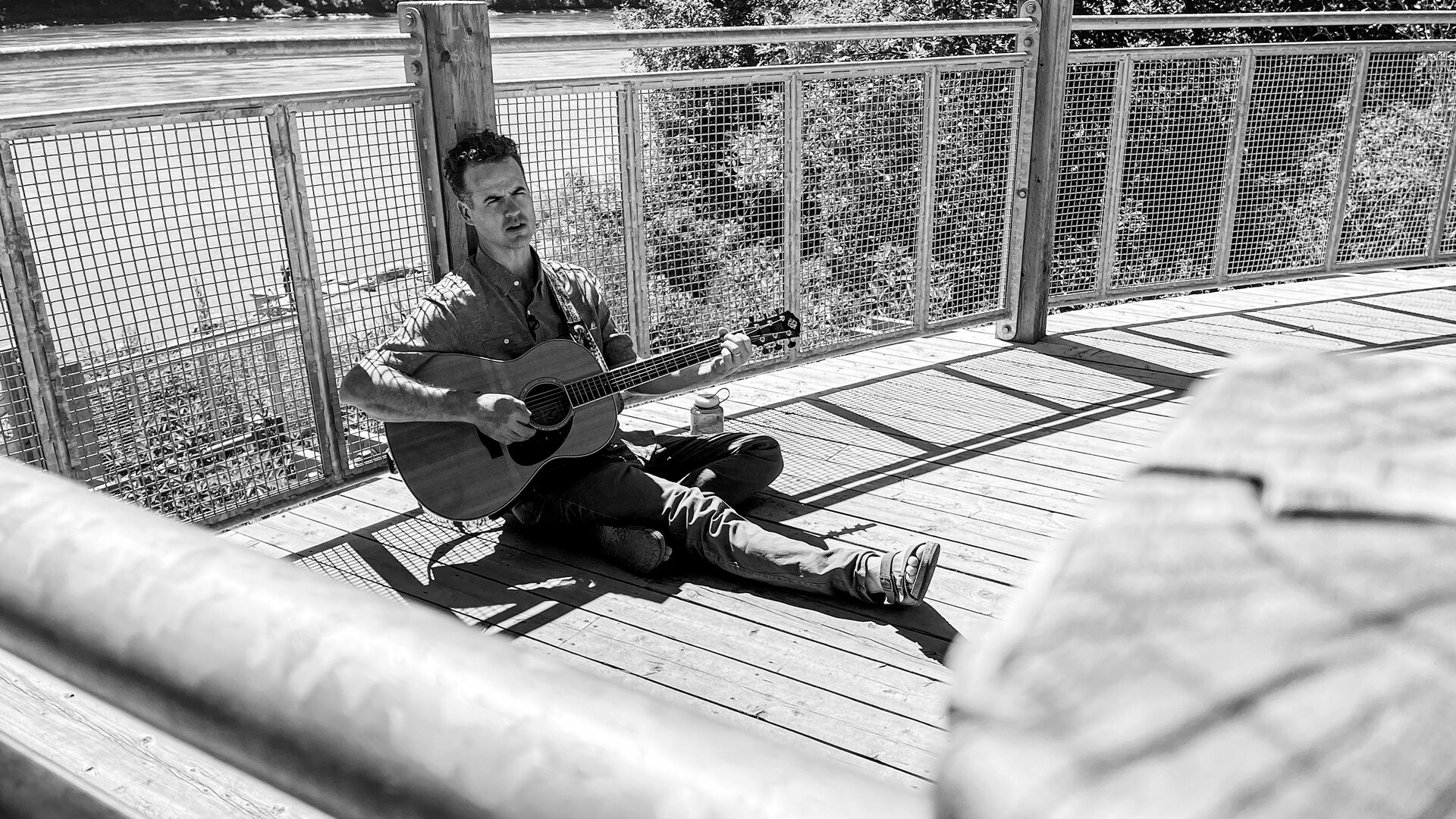 Un homme assis sur un balcon de bois, près d'une rivière, avec sa guitare