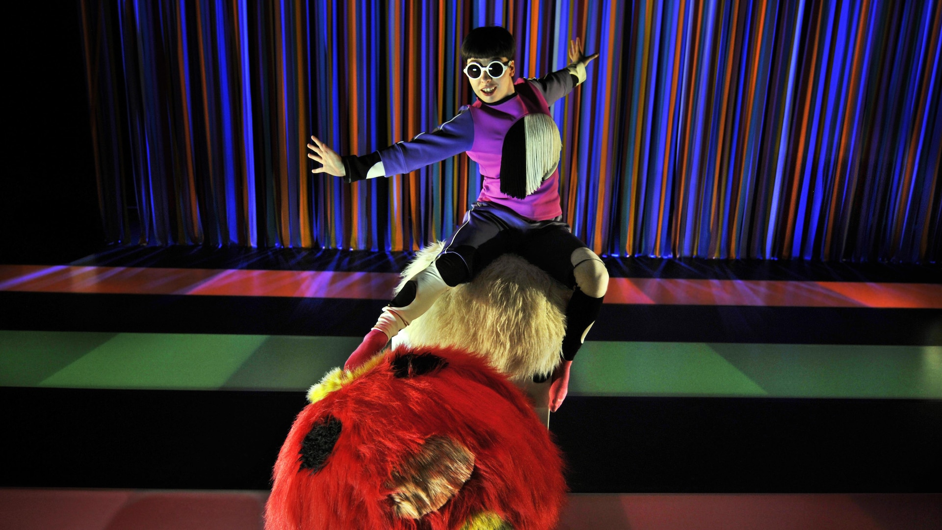 Une acrobate est assise, bras tendus, sur un gros rouleau de fourrure rouge et jaune.