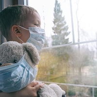 Un enfant portant un masque regarde par la fenêtre avec son toutou.