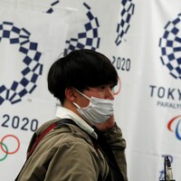 Un homme d'origine asiatique porte un masque sanitaire. Derrière lui, un bandeau fait la promotion des Jeux olympiques de Tokyo.
