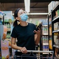Une femme asiatique portant un masque achète des aliments dans un supermarché. Elle tient son cellulaire dans une main. Elle regarde les prix des aliments sur les étagères. 