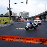 Une motocyclette est photographiée derrière un ruban de policier où il est écrit : « passage interdit ». Sur cette même rue, une voiture de police qui se trouve derrière la moto a ses gyrophares allumés.