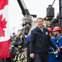 Andrew Scheer marche, souriant, près d'une rangée de travailleurs.