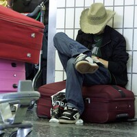 Un homme avec un chapeau de cowboy baisse la tête. Il est assis sur une valise, par terre dans un aéroport. Un chariot avec des valises empilées se trouve près de lui.