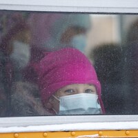Une enfant coiffée d'une tuque rose et portant un masque bleu sur le visage est assise dans un autobus scolaire et regarde par la fenêtre.
