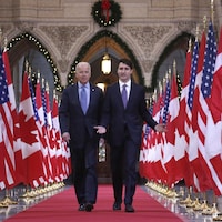 Joe Biden et Justin Trudeau marchent côte à côte au milieu d'une rangée de drapeaux canadiens et américains.