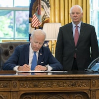Joe Biden signe la loi au bureau ovale de la Maison-Blanche devant un homme et une femme, qui sont debout.