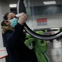 Un femme de profil portant un masque nettoie à l'aide d'une lingette l'intérieur d'une laveuse pour y déposes ses vêtements.