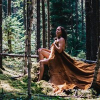 Une femme est assise sur un rocher en forêt. Sa peau nue est recouverte d'une large étoffe faite à partie d'une peau de gibier.