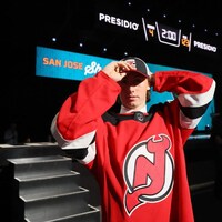 Cam Squires, vêtu d'un chandail des Devils, ajuste sa casquette. Derrière lui, l’estrade du repêchage de la LNH.