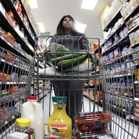 Une femme vue de face dans une épicerie. Elle pousse un chariot plein et avance entre deux rangées remplies de nourriture.