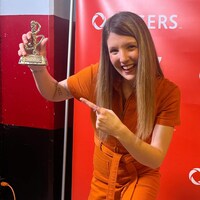 Chloé Breault debout et souriante, vêtue d'une combinaison orange, pointe le trophée qu'elle tient dans son autre main.