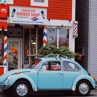 Une voiture Coccinelle de Vollkswagen, chargée d'un sapin de Noël sur le toit, garée devant un salon de barbier. 
