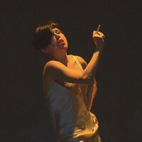 Une comédienne aux cheveux courts et vêtue d'une camisole blanche fume sur scène sous un éclairage sombre.