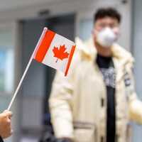 Une main agite un petit drapeau du Canada devant un homme avec un masque qui marche dans l'aéroport.