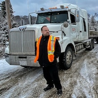Un homme barbu vêtu d'une veste de sécurité orange se tient devant un semi-remorque.