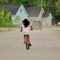 Une fillette sur son vélo au milieu d'une rue sur laquelle circule aussi une camionnette. 