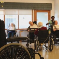 Au premier plan : un fauteuil roulant. Au fond, plusieurs personnes aussi en fauteuil roulant, dans un établissement de soins.
