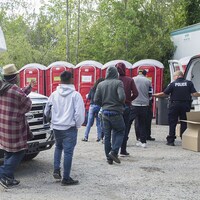 Six demandeurs d'asile arrivés par le chemin Roxham, près de Saint-Bernard-de-Lacolle, reçoivent des boîtes à lunch des mains d'un policier, alors qu'on voit des toilettes chimiques en arrière-plan.