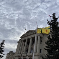 Le drapeau est hissé devant l'Assemblée législative de la Saskatchewan, à Regina. Le ciel est couvert avec quelques éclaircies en fond, et un sapin se tient à la droite du drapeau.