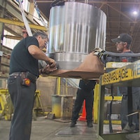 Des travailleurs manipulent une pièce mécanique suspendue dans une usine. 