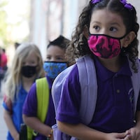 Des élèves en rang, portant des masques et leur sac-à-dos, marchent vers l'entrée de leur école.