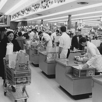 Une femme passe avec son panier d'épicerie rempli derrière la rangée de caisses d'un supermarché Steinberg.