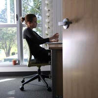 Assise à son bureau, une étudiante suit un cours en ligne de chez elle avec une fenêtre en arrière-plan.