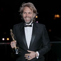 Un homme en smoking esquisse un large sourire en tenant un Oscar dans sa main. 