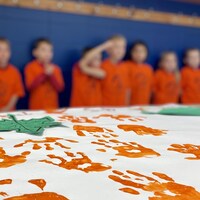 Une bannière contenant des traces de main orange est déposée sur une table. Derrière, on voit des silhouettes d'enfants floues.