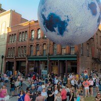 Une immense lune gonflable flotte au-dessus d'une foule dans une rue piétonnière en été.
