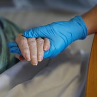 Une main ganté tient dans sa main une main d'une personne âgé couché dans un lit.
