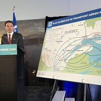 François Bonnardel parle au micro à côté d'une grande carte du Québec qui montre quelles destinations seront offertes.