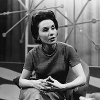 Dans un studio de télévision, la journaliste Françoise Faucher est assise sur une chaise, joignant ses mains devant elle.