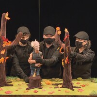 Sur une scène quatre personnes, toutes vêtues de noir et masquées, manipulent des marionnettes.