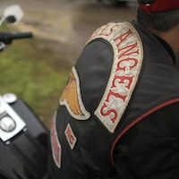 Les lettres des Hells Angels sont cousues au dos d'un manteau d'un homme  qui se tient près d'une motocyclette.