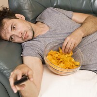 Un homme allongé sur un sofa mange des croustilles en regardant la télévision.