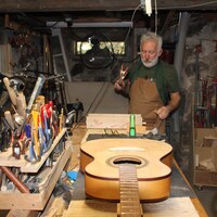 L'homme entouré d'outils et de planches de bois travaille dans un sous-sol. Une guitare est posée sur la table.