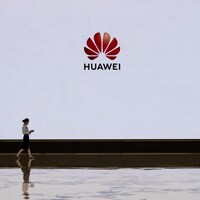 Une femme marche devant un écran blanc sur lequel est projeté le logo de la compagnie Huawei.