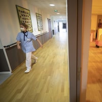 Une infirmière marche d'un bon pas dans un corridor. On voit une femme âgée dans un fauteuil roulant dans une pièce adjacente à ce couloir.