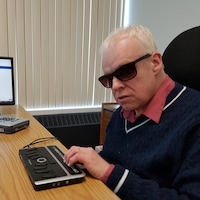 René Binet, une personne aveugle, est assis à un bureau. Il consulte un site internet à l'aide d'une plage braille.