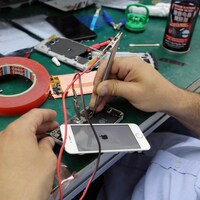 On voit les mains d'une personne qui manipule un iPhone à l'aide d'un outil sur une table de travail.