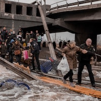 Des gens traversent une rivière sur une planche de bois sous un pont détruit.