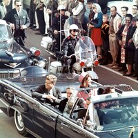 John F. Kennedy est assis à l'arrière d'une en voiture décapotable accompagné de son épouse lors d'un défilé, escorté par un policier à moto.