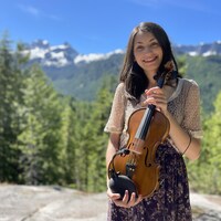 Jocelyn Pettit, souriant, tenant son violon face à la caméra. Elle prend la pose devant un paysage de montagnes.