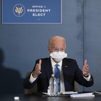 Joe Biden porte un énorme masque et est assis sous une pancarte disant : Bureau du président désigné.