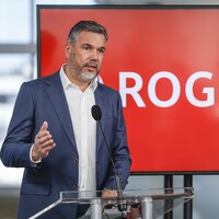 Jorge Fernandes parle dans un micro. Derrière lui se trouve une grande télévision qui affiche le logo de Rogers. La photo a été prise à Calgary le 28 avril 2022.