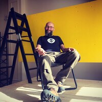 L'artiste José Luis Torres assis au coeur de son oeuvre, constituée d'une échelle noire et d'un panneau de bois peint en jaune. 